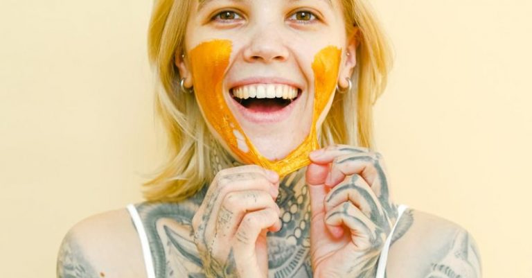Therapy - Young cheerful female with tattooed body and blond hair removing peel off golden mask from face looking at camera