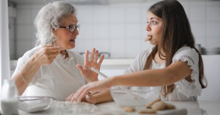 Others - Calm senior woman and teenage girl in casual clothes looking at each other and talking while eating cookies and cooking pastry in contemporary kitchen at home