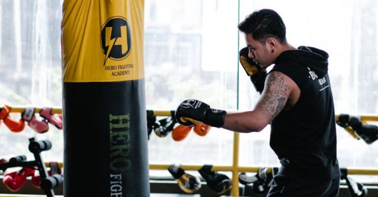 Techniques - Full length side view of barefoot Asian boxer in gloves punching bag during workout in light gym